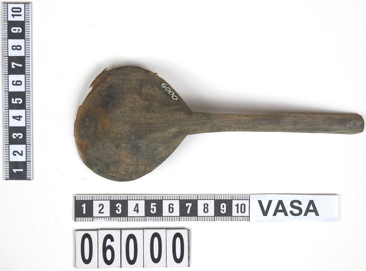 En träsked där skedbladets kant är skadad. Skeden har en mörkbrun ton på baksidan vilket kan tyda på bemålning. Skaftet är ovalt.