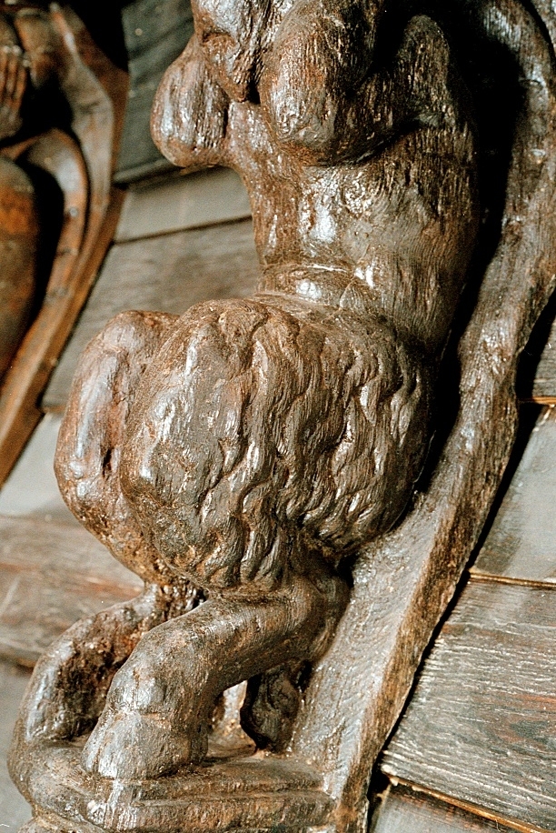 Skulptur föreställande en djävulsfigur i starkt hukande ställning, placerad på en hög sockel.
Figurens huvud är utrustat med stora, krumma horn. I vardera handen håller den på ömse sidor av huvudet något som möjligen kan tolkas som en stor snäcka eller lur. Hakan pryds av ett bockskägg. Figuren har fladdermusvingar på ryggen och starkt luden underkropp med klövförsedda bockfötter. Fötterna vilar på den starkt sluttande sockeln, som framtill är prydd med en ap- eller lejonliknande maskaron med stor nos och hög, välvd panna. På sockelns sidor ses voluter.
Sockeln utgör fortsättning på det kraftiga klink-konstruerade bakstycket som är framdraget ovanför figuren.
Skulpturen är delvis kraftigt sliten och vittrad, särskilt den övre delen. Nederdelen är något bättre bevarad.

Text in English: Sculpture of a devil figure in a crouched position, placed on a high socle.
The figure''s head is equipped with large, curved horns. In either hand the figure holds on either side of its head something that may possibly be identified as a large seashell or horn. The devil has a goat''s beard on its chin, bat wings on its back and very hirsute lower parts of the body, and cloven goats'' feet.
The feet rests on a sharply sloping socle that is decked in front with a mask of an ape-like or lion-like face with a large nose and a high arched forehead. On the sides of the socle volutes are visible.
The socle forms the extension of the heavy, clinker-built back-piece, which is drawn forward above the figure.
The sculpture, particularly its upper parts, is much worn and disintegrated. The lower part is somewhat better preserved.