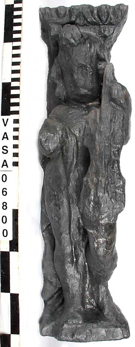 Skulptur föreställande en barnfigur spelande på en viola da braccio.
Fiolen hålles med vänster hand och stöder mot vänster knä. Kroppstyngden vilar på höger ben. Huvudet är vridet åt höger. Antydan till mantel utmed höger sida. Ovanför hjässan ses en äggstavslist. Figuren står på ett minimalt underlag som troligen avser att föreställa ett stycke naturlig mark. Skulpturens baksida är slät
Skulpturen är kraftigt sliten. Höger underarm är bortbruten.

Text in English: This is a sculpture of a child holding a viola da braccio in the left hand and supported on the left knee.
The weight of the body rests on the right leg. The head is turned to the right. There is the suggestion of a mantle on the figures right side. Just above the head, the back board comes forward at right angles and the front of the board is decorated with an egg moulding design. The figure stands upon a small plinth. The back of the sculpture is smooth.
The sculpture is very worn and the right forearm is broken off.