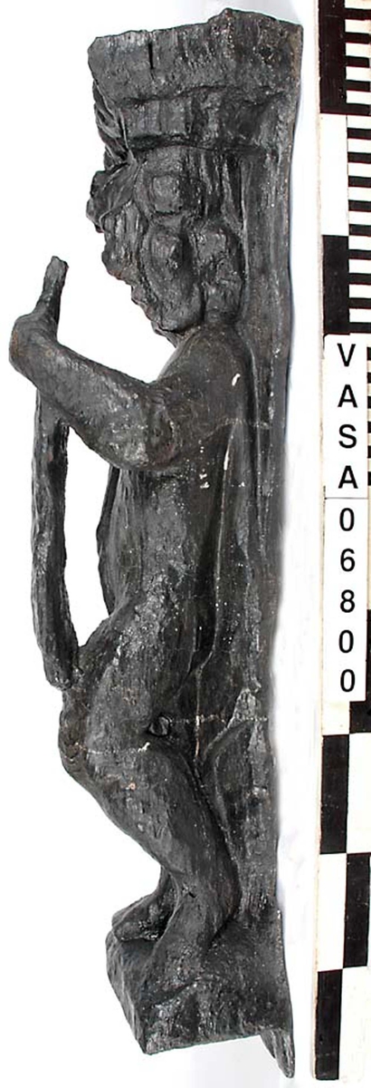 Skulptur föreställande en barnfigur spelande på en viola da braccio.
Fiolen hålles med vänster hand och stöder mot vänster knä. Kroppstyngden vilar på höger ben. Huvudet är vridet åt höger. Antydan till mantel utmed höger sida. Ovanför hjässan ses en äggstavslist. Figuren står på ett minimalt underlag som troligen avser att föreställa ett stycke naturlig mark. Skulpturens baksida är slät
Skulpturen är kraftigt sliten. Höger underarm är bortbruten.

Text in English: This is a sculpture of a child holding a viola da braccio in the left hand and supported on the left knee.
The weight of the body rests on the right leg. The head is turned to the right. There is the suggestion of a mantle on the figures right side. Just above the head, the back board comes forward at right angles and the front of the board is decorated with an egg moulding design. The figure stands upon a small plinth. The back of the sculpture is smooth.
The sculpture is very worn and the right forearm is broken off.