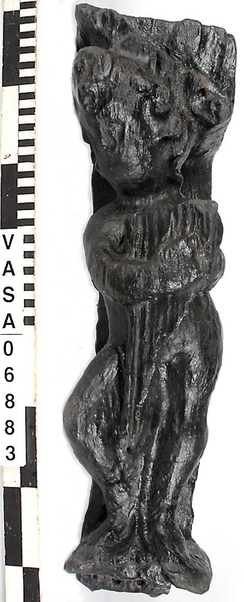 Skulptur föreställande en barnfigur spelande en harpa.
Instrumentet hålls med bägge händerna. Huvudet är vridet något åt höger. Kroppstyngden vilar på vänster ben medan höger ben är böjt. Vid höger sida syns en mantel. Ovanför hjässan sitter en snedställd äggstavslist. Figuren står på ett minimalt underlag som troligen avser att föreställa ett stycke naturlig mark. Skulpturens baksida är slät.
Skulpturen är mycket sliten.

Text in English: This is a sculpture of a child playing a harp.
The instrument is held in both hands. The head is turned slightly to the right. The weight of the body rests on the left leg, the right leg is bent. A mantle is to be seen to the figures right side. Just above the head the back board comes foreward at right angles and the front of the board is wide and is decorated with an egg moulding design which is placed diagonally across it with the right-hand side lower than the left-hand side, when the sculpture is viewed from the front. The figure stands upon a small flat plinth. The back of the sculpture is smooth.
The sculpture is very  worn.