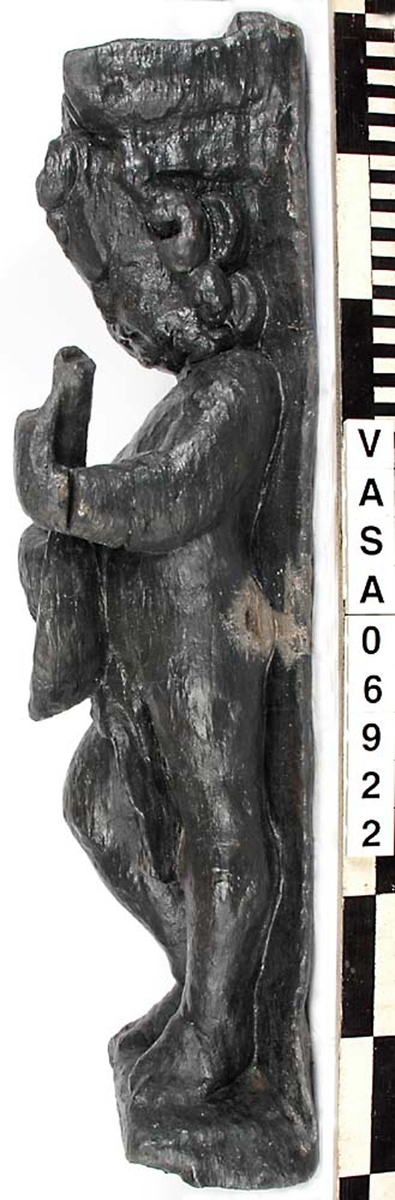 Skulptur föreställande en barnfigur med en cister (ett stränginstrument som tillhör familjen lutor).

Instrumentet hålls med bägge händer framför kroppen. Huvudet något vridet åt vänster. Kroppstyngden vilar på vänster ben medan höger ben är böjt. Utmed höger sida skymtar en mantel. Figuren står på ett minimalt underlag som troligen avser att föreställa ett stycke naturlig mark. Skulpturens baksida är slät.

Skulpturen är välbevarad.



Text in English: This is a sculpture of a child playing a cister.

It holds the instrument in both hands in front of its body. The head is turned slightly to the left. The weight of the body rests on the left leg, the right leg is bent. A mantle or cloak is to be seen on the figures right side. The child stands upon a small flat plinth. The back of the sculpture is smooth.

The sculpture is well preserved.