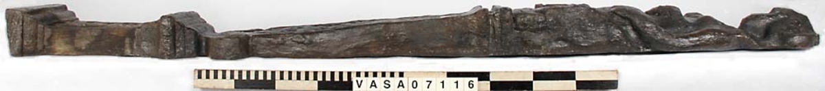 Skulpterad pilaster med övre delen i form av en karyatid med yppig, konstrik frisyr.
Höger arm hålls böjd med handen placerad framför bröstet. Vänster arm hålls utsträckt utefter sidan med handen vilande mot höften. Underkroppen döljs av ett hölje som framtill är prytt med stora, spiralformade ornament och små djurfigurer.
Karyatiden avgränsas från det rullverksornerade pilasterskaftet av en profilerad list. Nedtill avslutas skaftet av ett rullverksornament ovanför den rikt profilerade basen. Nedtill är pilastern försedd med en hög piedestal, prydd med beslagsornamentik. Figuren och dekoren omfattar även pilasterns sidor. Baksidan är slät.


Text in English: Sculpted pilaster with its upper part shaped as a Caryatid with a luxuriant artistic coiffure.
The right arm is held bent with the hand in front of the breast, the left arm is held at the side with the hand resting against the thigh. The lower part of the body is concealed by a shift or slip that is decorated with large spiral-shaped ornaments and small figures of animals.
A profiled batten separates the Caryatid from the shaft of the pilaster, which is ornamented with scrollwork. The shaft terminates with a scrollwork ornament above the richly profiled base. Below, the pilster is equipped with a high pedestal, decked with applied ornamentation. The back is smooth.