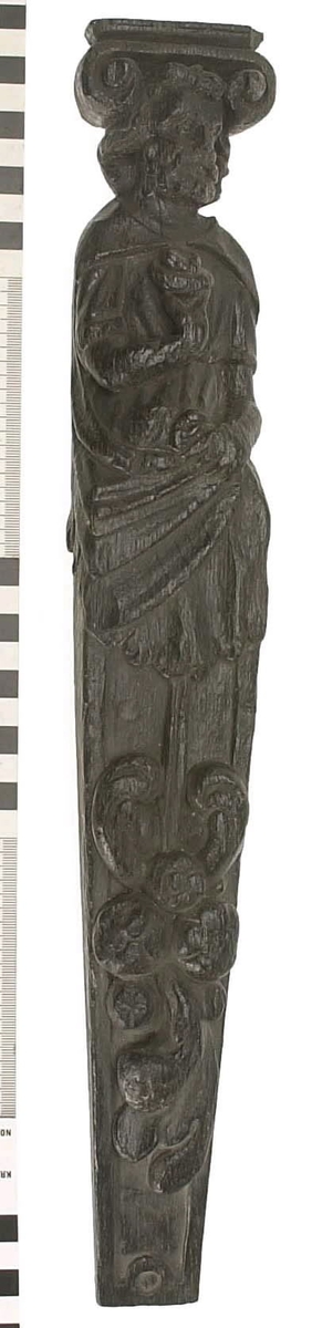 Hermpilaster.
Liten skulpterad pilaster föreställande skäggig herm under joniskt kapitäl.
Huvudet är vridet mot vänster. Hermen är insvept i en mantel som hålls fast med vänster hand framför magen. I höger hand håller figuren ett okänt föremål (blomsterkälk?) framför bröstet. Pilasterskaftet är prytt med stora frukter och blad, fasthängande vid en stängel som delvis täcker det nedåt avsmalnande skaftets ramverk. Baksidan är slät.
Skulpturen är mycket välbevarad med sju spikhål.

Text in English: Small sculpted pilaster with a bearded Herm beneath an Ionic capital.
The head of the figure is turned to the left. The Herm is enveloped in a mantle, held fast with the left hand in front of its stomach. Its right hand holds an unknown object (a bunch of flowers?) in front of its breast. The shaft of the pilaster is decked with large fruit and leaves hanging from a stalk which partly cover the framing of the tapering shaft. Its back is flat.
The sculpture is very well preserved.