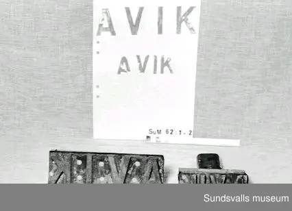 Stämpel i gummi med handtag i trä. Stämpeln visar bokstäverna AVIK. Röda färgrester finns kvar på stämpeln.