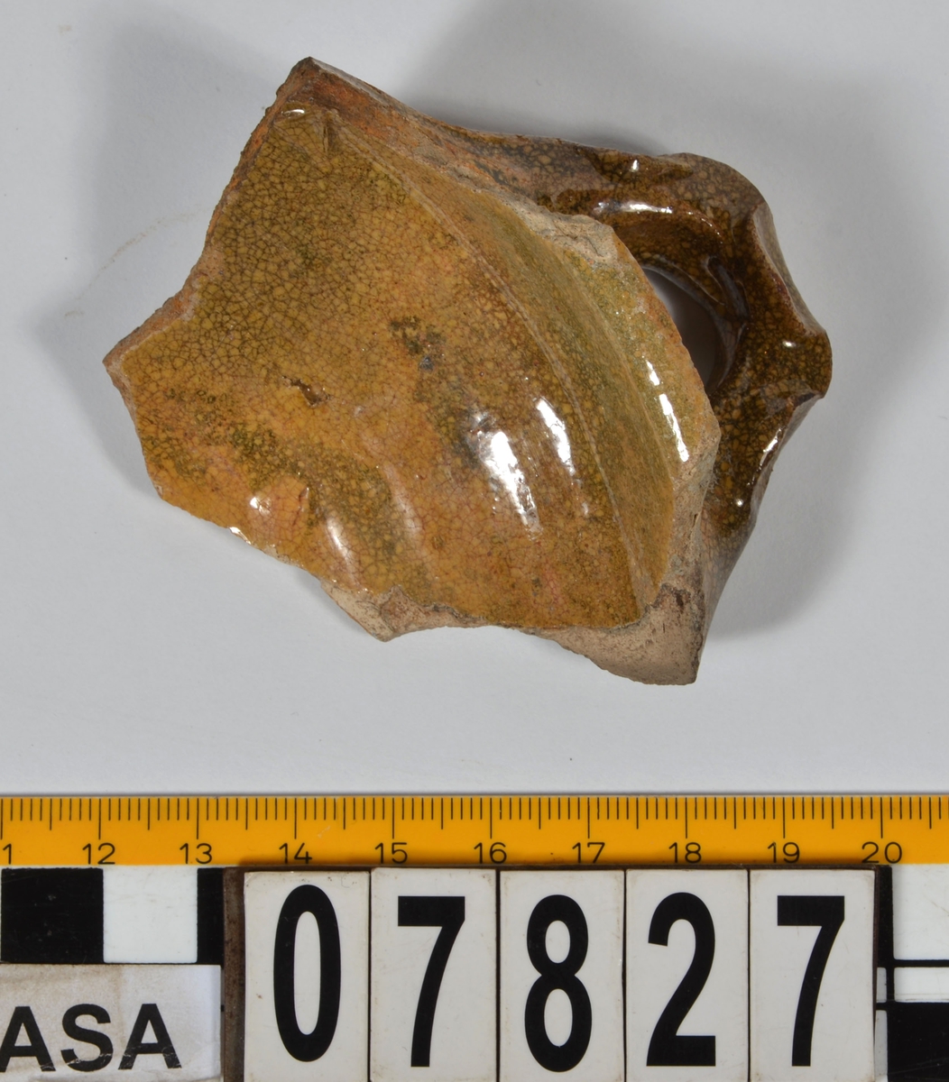 Del av skål.
1 st vertikal bandhänkel, tummad, 70 x 60 mm. Fragment av mynningsrand och buk. In- och utsida blyglaserad gulgrön.