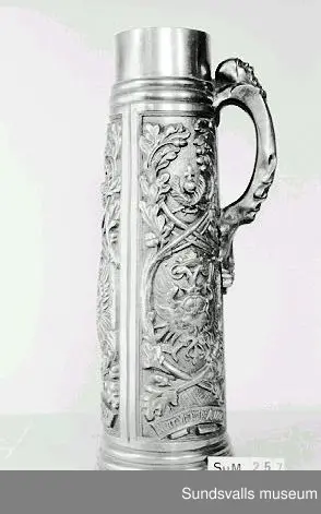 Dryckesstop i tenn med gjuten dekor i form av ett vapen med en örn, grenar, blad och ekollon. Reliefdekor på hänkeln.