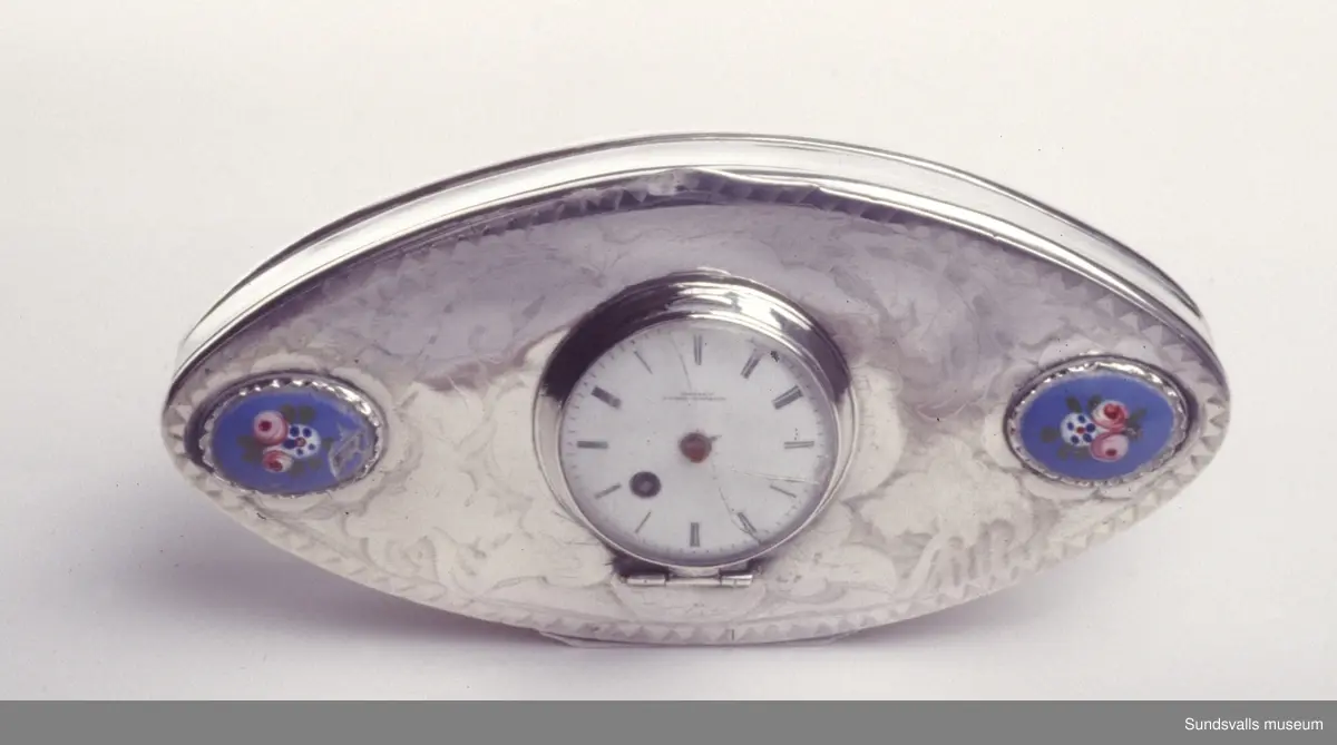 Snusdosa i silver. Spetsoval med profilerade sidor. På locket svisselerad dekor, samt två infattade emaljmålningar och ett infattat nyckeldragarur. Uret är märkt 'Vacheron Freres' på verket och tavlan.