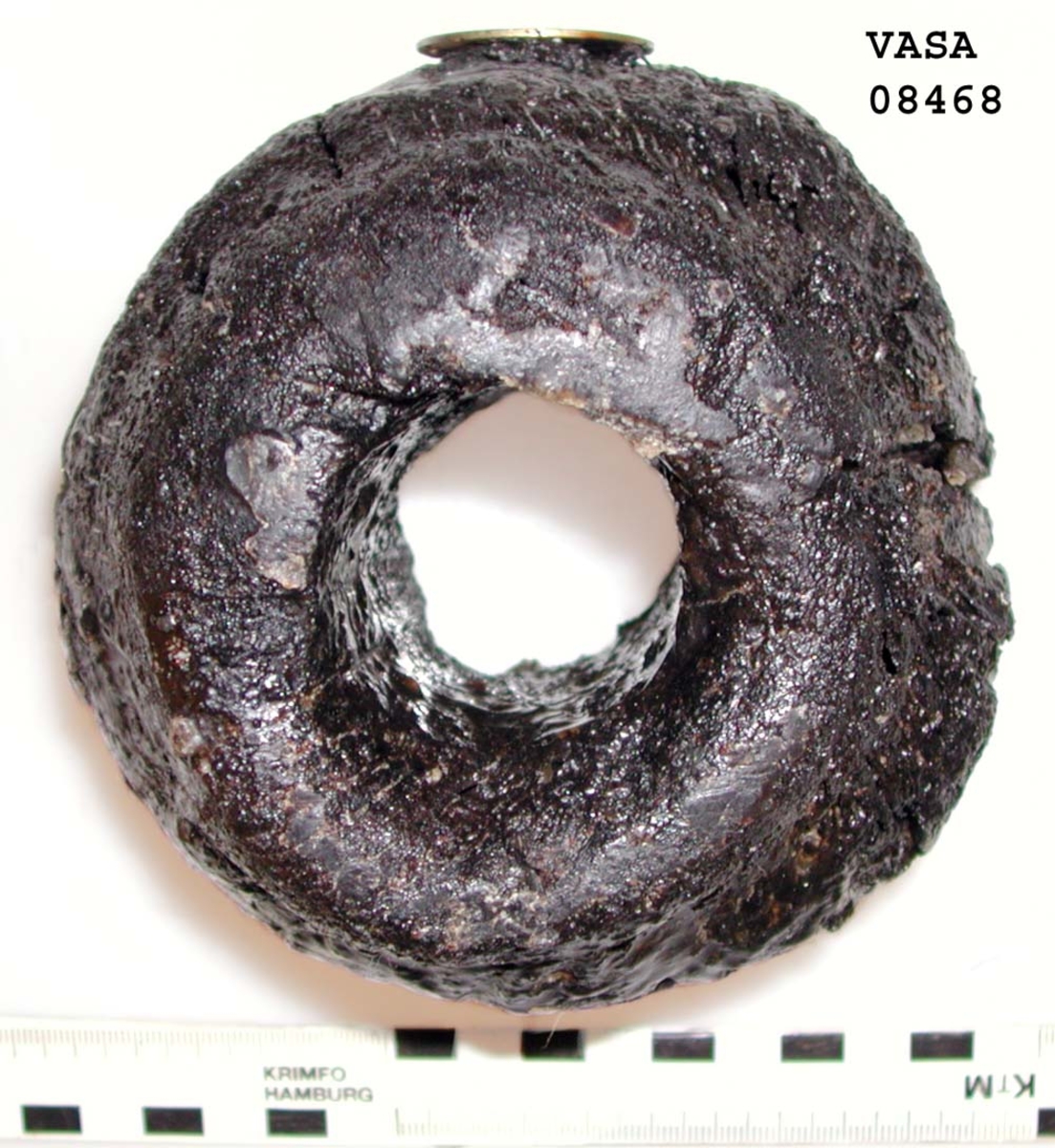 Kolv till ansättare. Kolven består av ett cylindriskt svarvat trästycke med ett centrumhål för skaftet. I kolvens övre ände, mot skaftet till, är kolven profilsvarvad. Kolven är kraftigt eroderad och sprucken. Profilsvarvningen upptill är nedsliten.