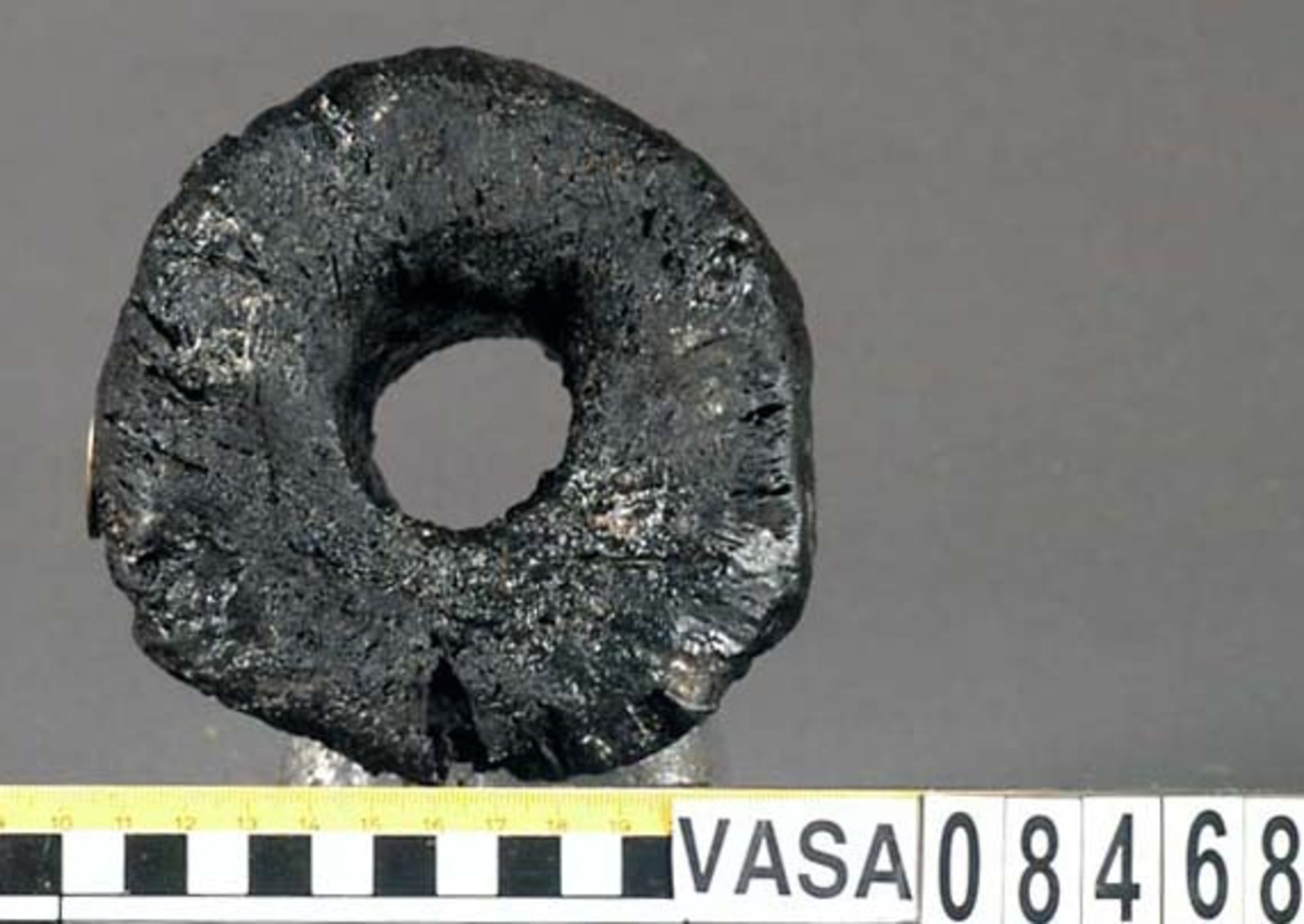 Kolv till ansättare. Kolven består av ett cylindriskt svarvat trästycke med ett centrumhål för skaftet. I kolvens övre ände, mot skaftet till, är kolven profilsvarvad. Kolven är kraftigt eroderad och sprucken. Profilsvarvningen upptill är nedsliten.