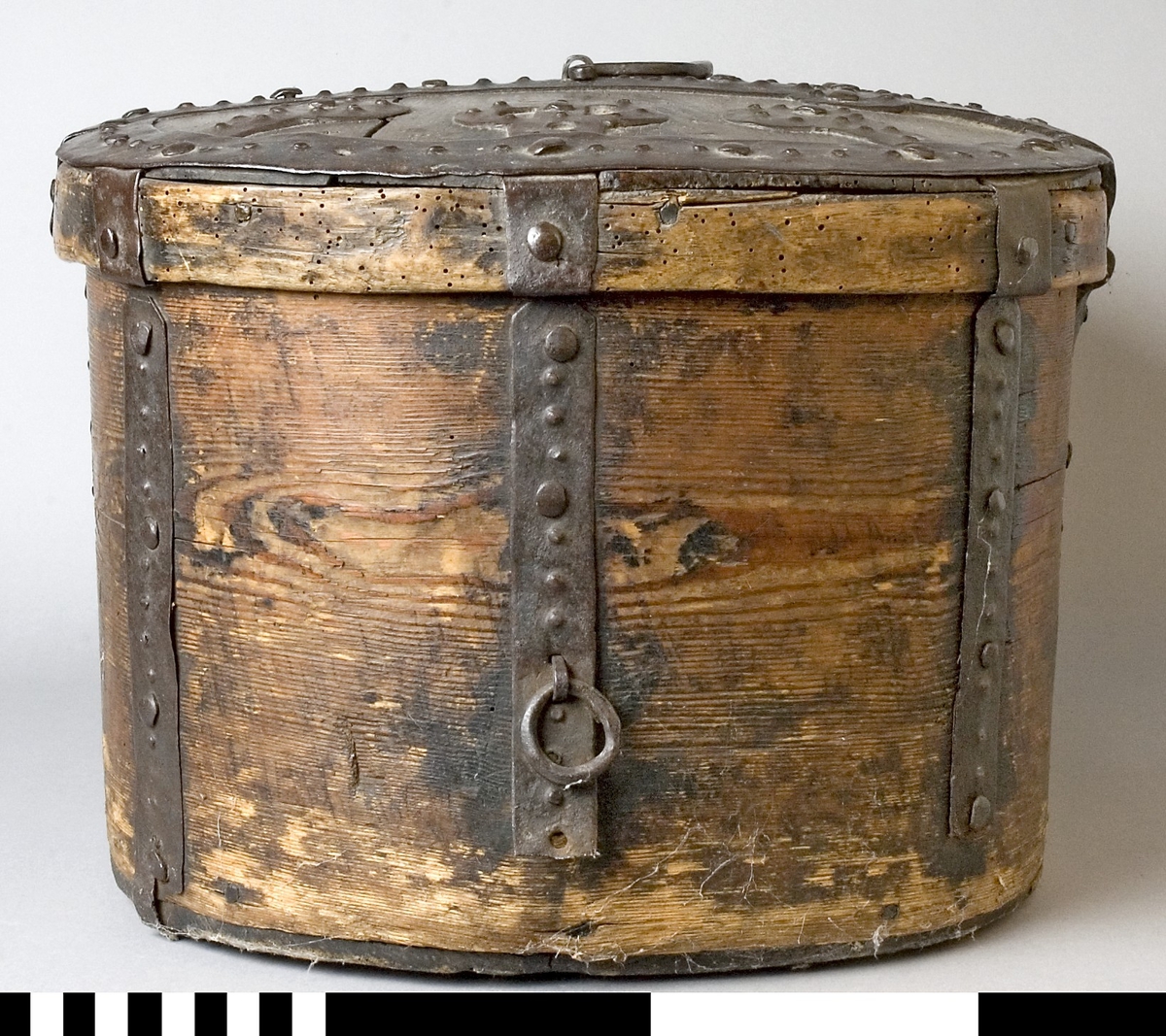 Färdskrin av trä med beslag av järn. Oval form med ett lock som är dekorerat med smida järnbeslag. Kanten på locket är skod med järn. På framsidan löper beslag ned och runt om. Centrerat mitt fram finns ett lås med nyckelskylt i järn.