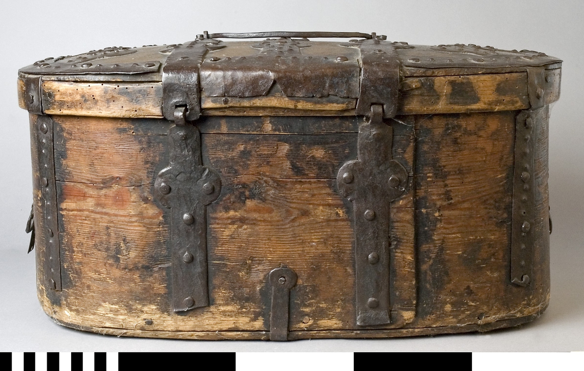 Färdskrin av trä med beslag av järn. Oval form med ett lock som är dekorerat med smida järnbeslag. Kanten på locket är skod med järn. På framsidan löper beslag ned och runt om. Centrerat mitt fram finns ett lås med nyckelskylt i järn.