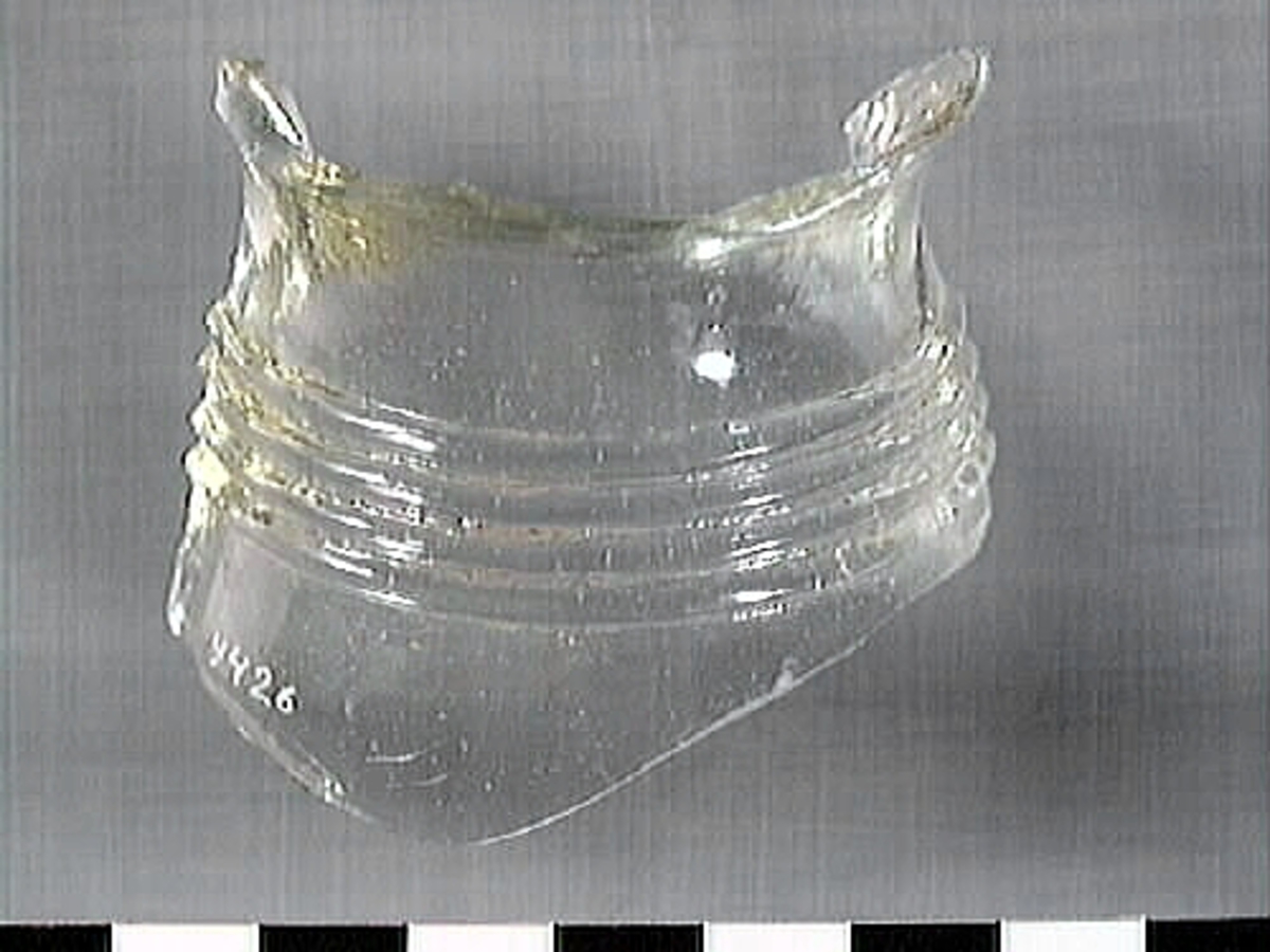 En glasskärva. Troligtvis härrör skärvan från ett dricksglas, eventuellt en flaska. Skärvan är försedd med en banddekor som består av tre glassträngar. Glaset är transparant och svagt grönfärgat.

2015: No damage. Glass is clear with a greenish.  Glass has three bands running horizontally.