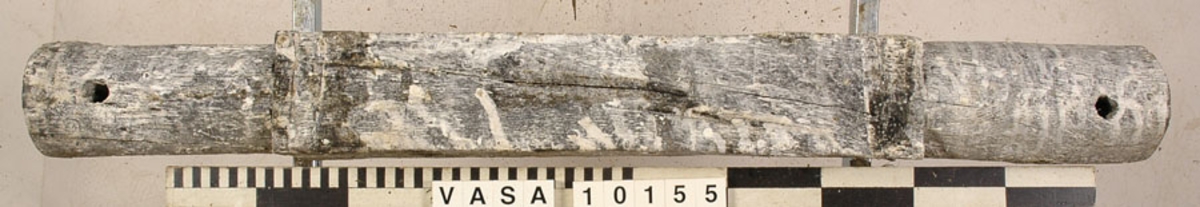 Lavettaxel, främre. Ett avlångt trästycke med kvadratiskt tvärsnitt mitt på samt cylinderformade ändar. Axeln var fastsatt i lavettbottnens undersida med två grova spikar och två järnband.

För mer information se Dokumentationsblankett under Referenser – Länkade filer.

Text in English: Axle (front) for gun carriage 10153, consisting of a square-section beam with round stub axles at the ends, originally fixed to a notch in the underside of the carriage bed by two nails and two iron straps.

For more information see the Documentation form under References – Linked files.