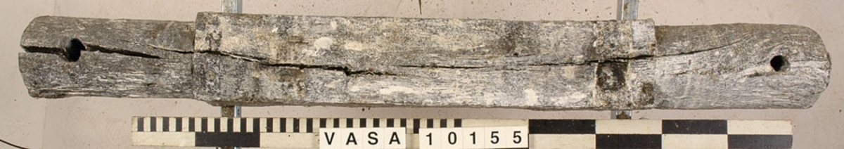 Lavettaxel, främre. Ett avlångt trästycke med kvadratiskt tvärsnitt mitt på samt cylinderformade ändar. Axeln var fastsatt i lavettbottnens undersida med två grova spikar och två järnband.

För mer information se Dokumentationsblankett under Referenser – Länkade filer.

Text in English: Axle (front) for gun carriage 10153, consisting of a square-section beam with round stub axles at the ends, originally fixed to a notch in the underside of the carriage bed by two nails and two iron straps.

For more information see the Documentation form under References – Linked files.