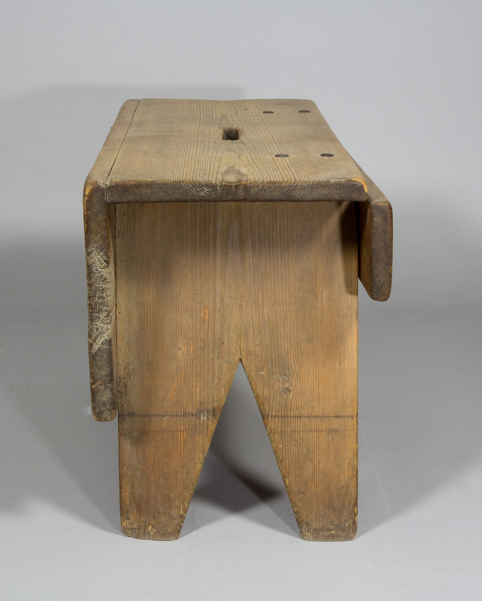 Pall eller pallbol i furu. Låg, avlång med bockformade ben. På pallens ena långsida väl tilltagen sarg, på andra sidan av fälltyp, fäst med gångjärn. Handtag i sätet i form av ett hål.