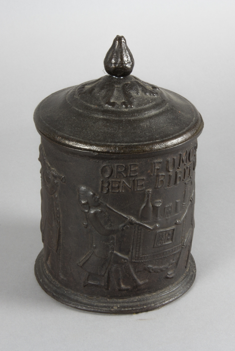 Relief på burken med text. Relief med rökande och spelande män. Några av männen sitter vid en spis. Riksvapen med flankerande lejon. Årtal 1794.
Text på burken. ORE FUMOSO BENE BIBENTUR.