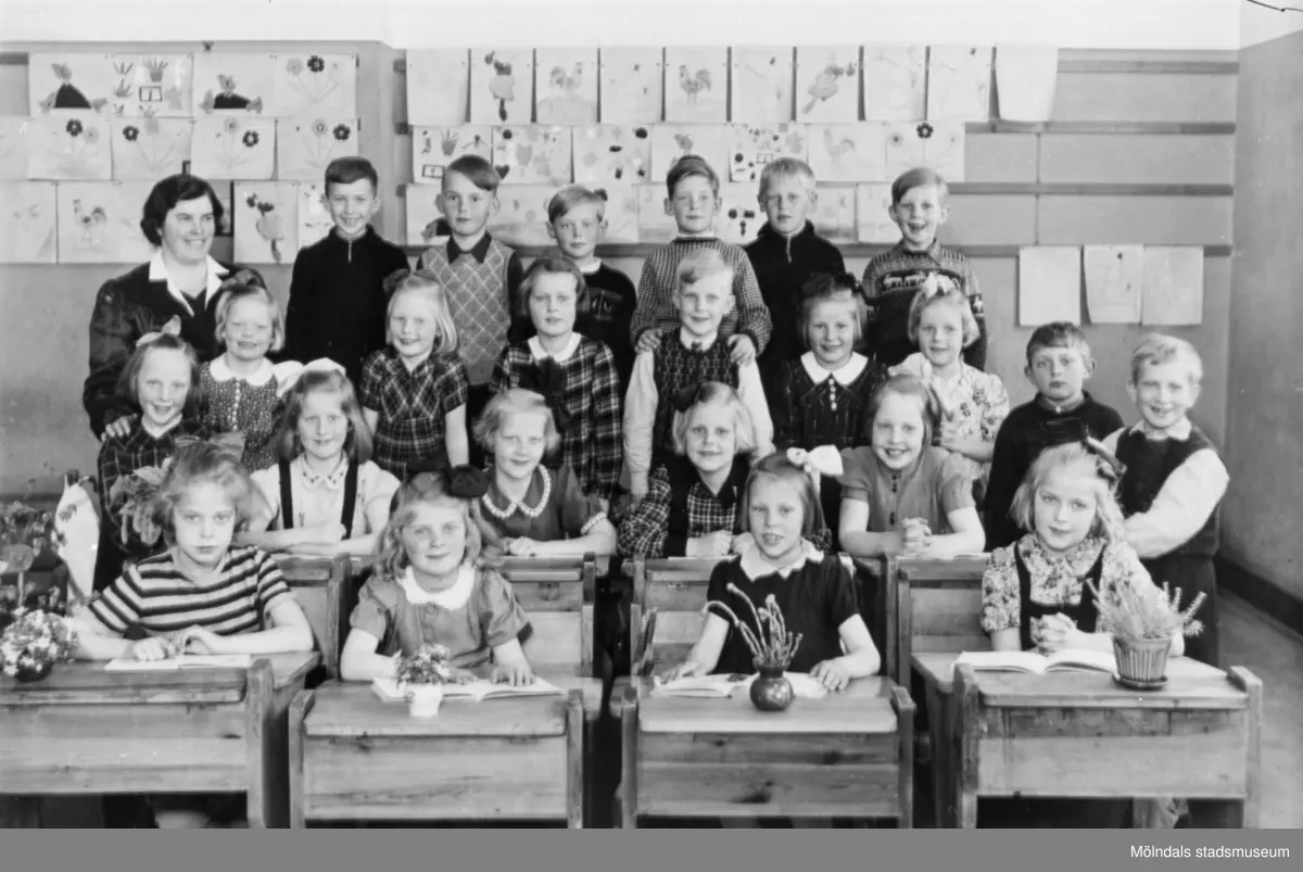 Fröken R. Sylvén med 1:a klass-elever på Centralskolan 1943-44. Flickor sittandes i bänkar. Flickor och pojkar ståendes bakom. Teckningar på väggen. Med på bild finns, sittandes till höger, Berit Gustafsson (född Larsson 1936).