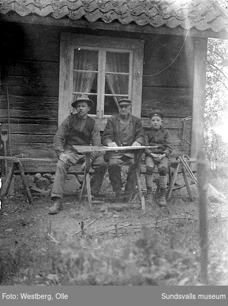 Två män och en pojke vid trädgårdsbord utanför ett bostadshus.