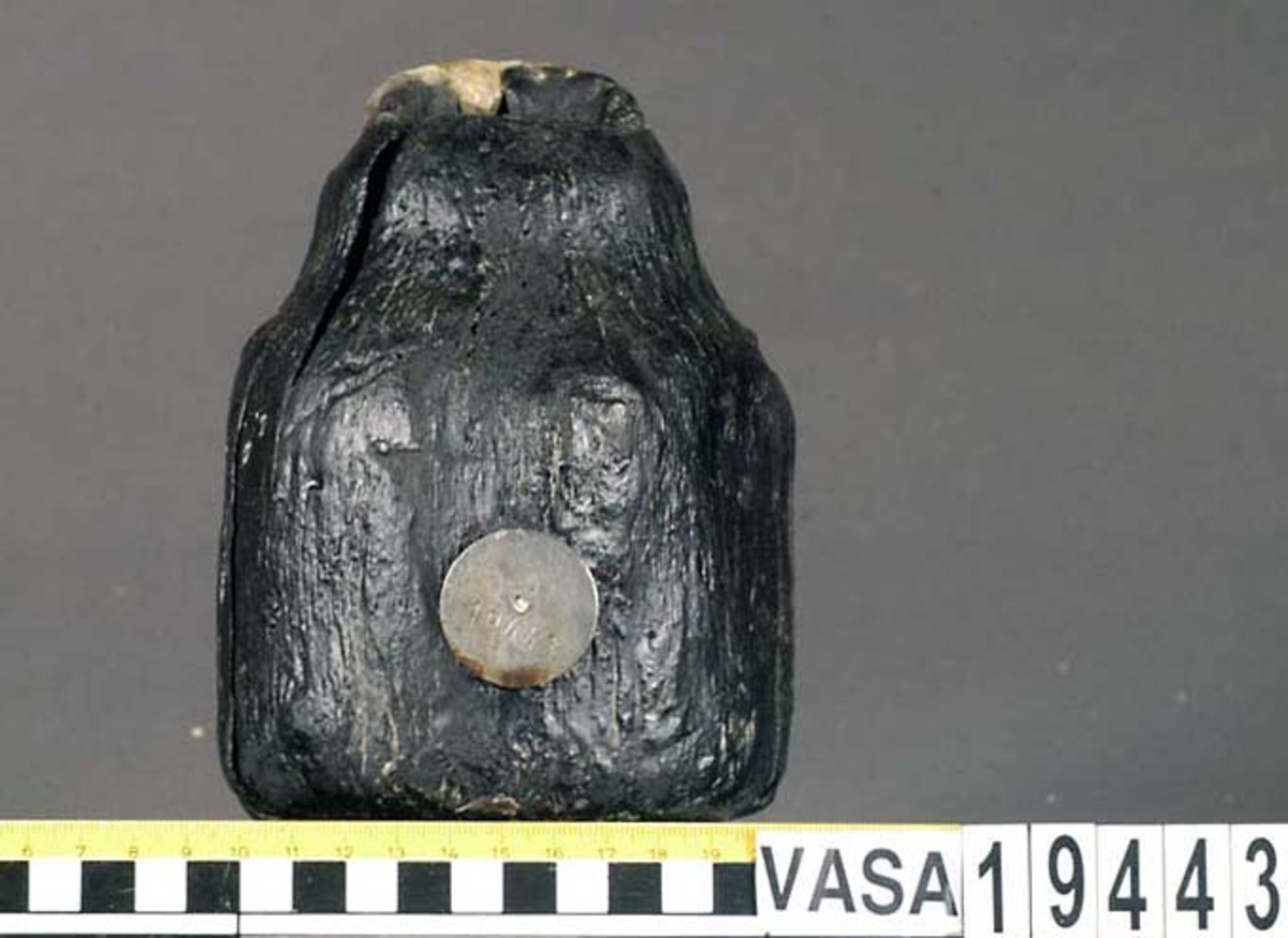 Kolv till ansättare. Kolven består av ett cylindriskt, svarvat, trästycke med ett centrerat skafthål. I övre änden, åt skaftet till, är kolven profilsvarvad. Kolven är kraftigt eroderad, mest på ena sidan. En kraftig spricka från utsidan in till skafthålet.