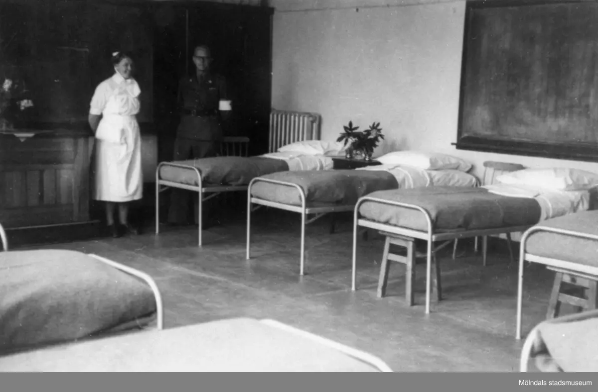 "Karantänsförläggning" på Kvarnbyskolan för överlevande från koncentrationsläger i Tyskland och Polen 1945.  
Skolan fungerade under denna sommar som beredsskapssjukhus.
 
En sjuksal med sängar samt en sköterska och en man från Röda Korset.
Den svarta griffeltavlan bakom sängarna till höger visar på att det är en av skolsalarna som gjorts om till sjukhussal.