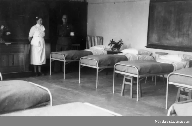 "Karantänsförläggning" på Kvarnbyskolan för överlevande från koncentrationsläger i Tyskland och Polen 1945.  
Skolan fungerade under denna sommar som beredsskapssjukhus.
 
En sjuksal med sängar samt en sköterska och en man från Röda Korset.
Den svarta griffeltavlan bakom sängarna till höger visar på att det är en av skolsalarna som gjorts om till sjukhussal.