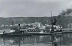 Sundsvalls hamn med båtar och magasin. I bakgrunden Norrmalm och Sundsvalls lasarett. Vykort.