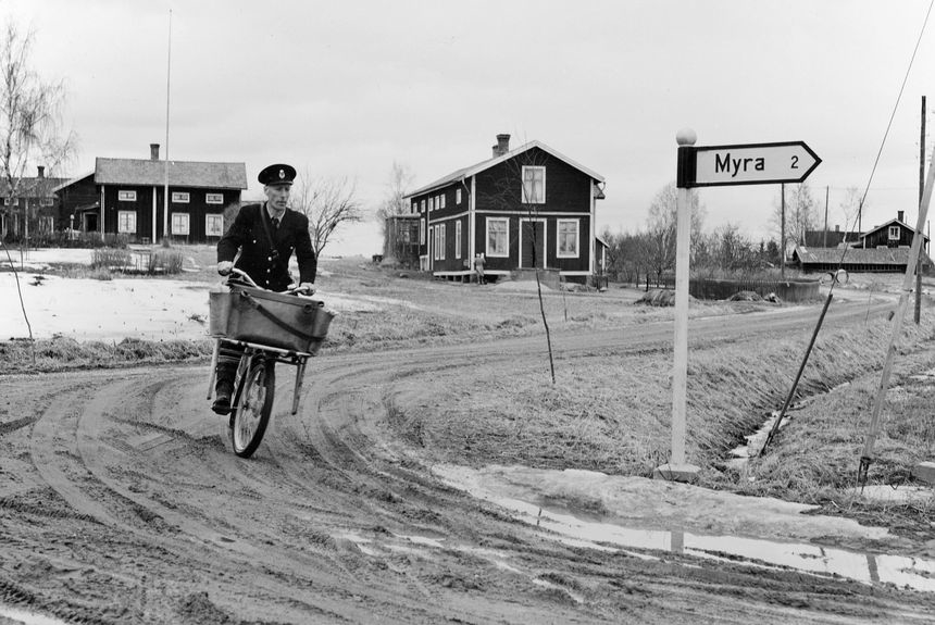 Lantbrevbärare, postiljon Erik Bergman (cykelåkande) i
Delsbo.  April 1956.

Brevbäringsturen är 2 ½ mil lång och omfattar nästan 300 hushåll. Postväskan väger ofta 30-40 kg.