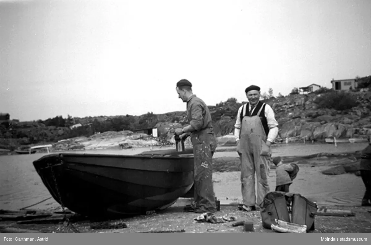 Pappa Helmer Garthman målar båten. I bakgrunden syns sönerna Alf och Leif som leker vid vattnet. Mannen i mitten är okänd av givaren. Näset, 1950-tal.