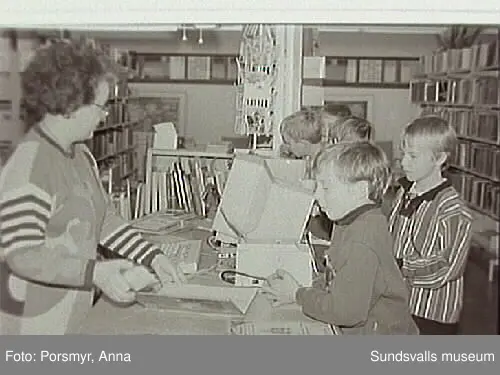 Dokumentationsfotografering av verksamheten vid biblioteksfilialen i Matfors. Bild 01Biblioteksassistenten Ingrid Engström, Matfors bibliotek, med låntagare vid utlånedisken. Bild 09 bibliotekaren Inger Barkman läser för en grupp förskolebarn i bibliotekets "läsrum".