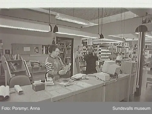 Dokumentationsfotografering av verksamheten vid biblioteksfilialen i Matfors. Bild 01Biblioteksassistenten Ingrid Engström, Matfors bibliotek, med låntagare vid utlånedisken. Bild 09 bibliotekaren Inger Barkman läser för en grupp förskolebarn i bibliotekets "läsrum".