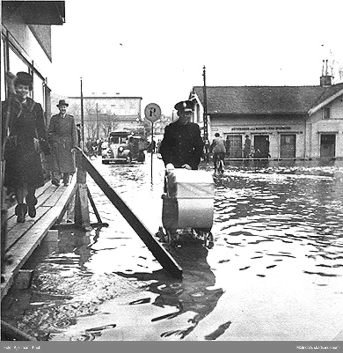 Polisman skjuter barnvagn på Frölundagatan under översvämning, 1940-tal. Från vänster ses gaveln på Frölundagatan 2, Kvarnbygatan 1 (höghuset i bakgrunden) och Kungsbackavägen 1.