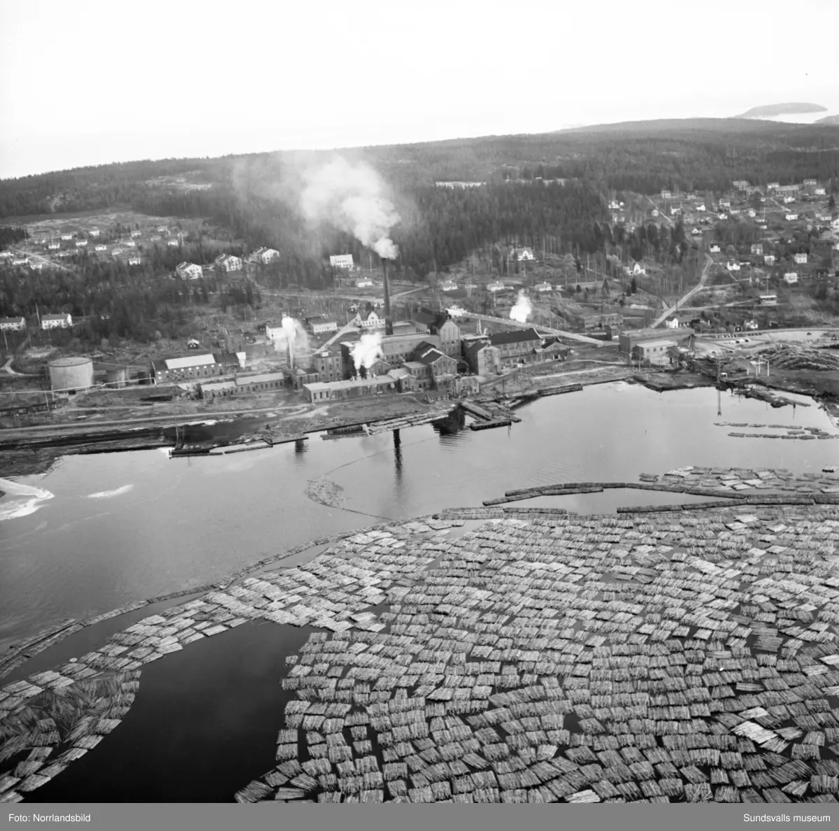 Flygfoton över Essvik och Nyhamn med timmerupplag och fabriksbyggnader.