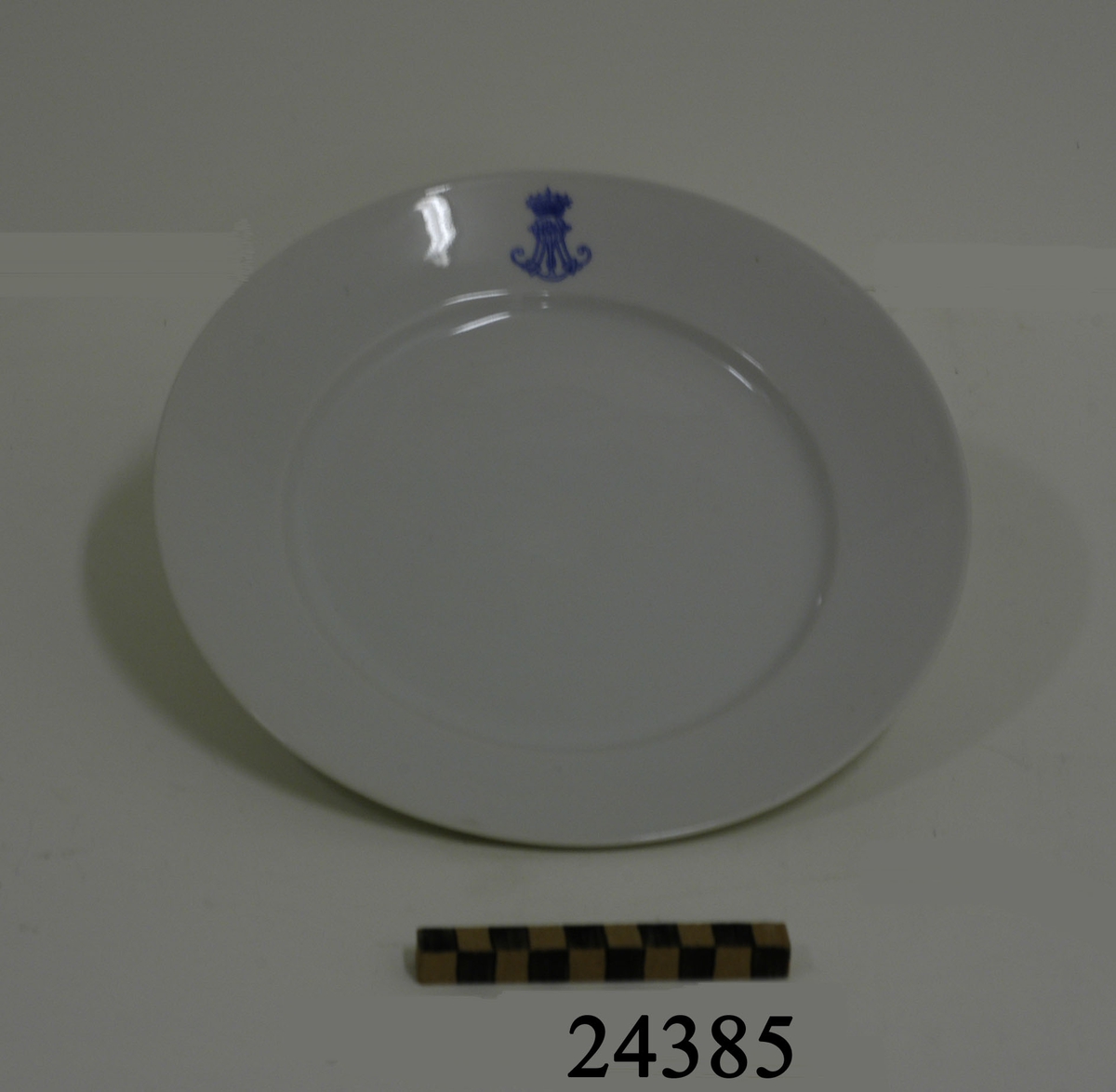 Flat tallrik av vitt porslin, glaserad, bräm bredd = 40 mm. På brämet ett blått emblem: ett krönt stockankare med monogram R M ( Reale Marina = Kungliga Flottan ). På undersidan fotring samt grön stämpel: " RICHARD GINORI 33 - 4. W 22.