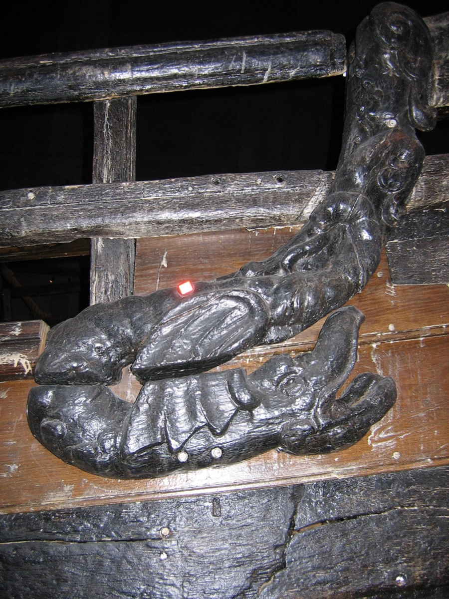 Skulptur av en triton, svängd mot höger.
Tritonen är framställd i vänster profil och avtecknar sig i hög relief mot det släta bakstycket. Tritonen har en stor näsa och treflikig mustasch. På huvudet sitter en hjälm med dekorativa bladvoluter. Armarna är ersatta av långa, flikiga bladslingor. Bålens övergång i den fjällförsedda fiskstjärten markeras av stora bladbildningar. Tritonens stjärt döljs bakom stjärten och vingen till en under liggande drake, se fyndnummer 21277. På baksidan finns urtag för relingslisten.
Skulpturen är mycket välbevarad.

Text in English: A sculpture of a Triton curved to the right.
The figure is seen in left profile, in high relief against a plain back board. The face has a large nose and a moustache that splits into three parts. On its head is a helmet with decorative leaf volutes. The arms are replaced by long, lobed leaf formations. The transition area between the male torso and the scaled fish-tail is decorated with large leaves. The rear is hidden behind a dragon''s coiled tail and broad wing situated below, see No. 21277. There are notches on the back that fit into the railing.
The sculpture is very well preserved.