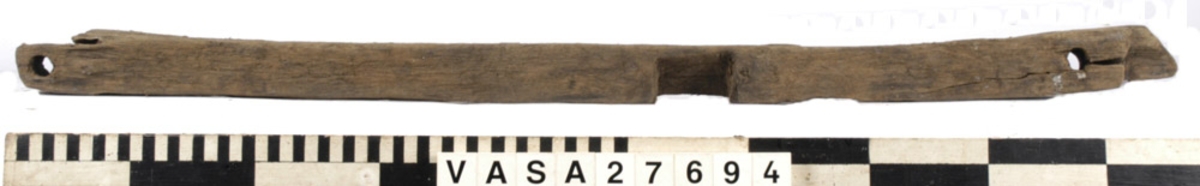 Relingslist. Den ena kortänden är uppfläkt. Den andra kortänden avslutas med en snedfasning, sannolikt en lask. Den andra slutar tvärt i sambands med en ursparning (sannolikt platsen för en årtull). Tolv stycken ej genomgående spikar i vad som sannolikt är den sida som vetter mot bordläggningen indikerar att den har spikats fast utifrån. Ett kraftigt genomgående hål syns i anslutning till ursparningarna för årtullarna. Spikhål syns i dessa avtrycks hörnor. På den sidan som vänder mot bordsidan och på undersidan syns snedfasade urtag, sannolikt för topparna till intimmer. I dessa urtagningar kan man skönja tydliga verktygsspår.