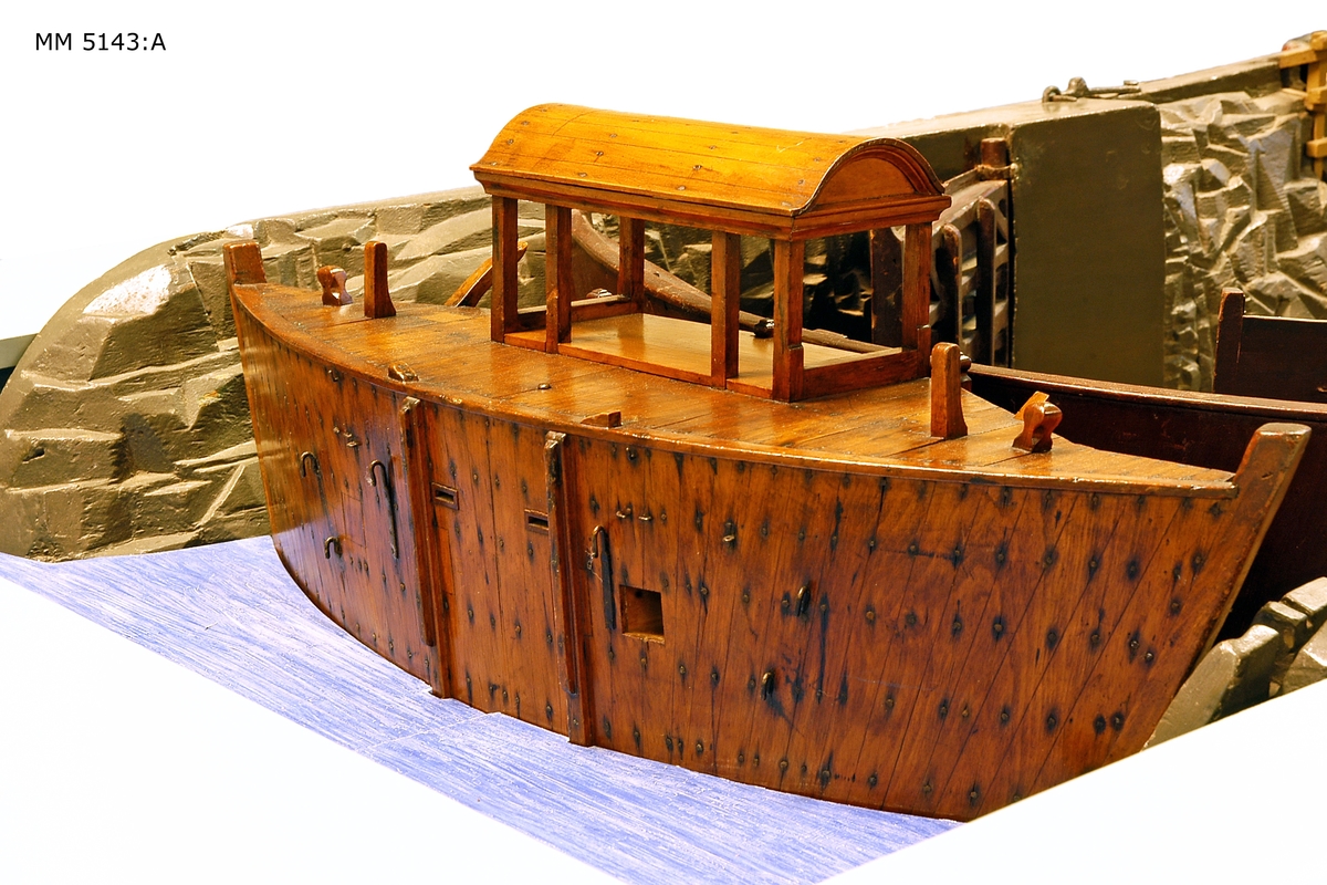 Modell av den s.k. Gamla dockan, eller Polhemsdockan på Lindholmen å Karlskrona örlogsvarv. Modellen försedd med sätt och lyftanordning för denna. Modellen tillverkad i två delar. Den är målad i mörkgrått, portarna fernissade. Blockraden i botten och trappan ned av omålat trä.