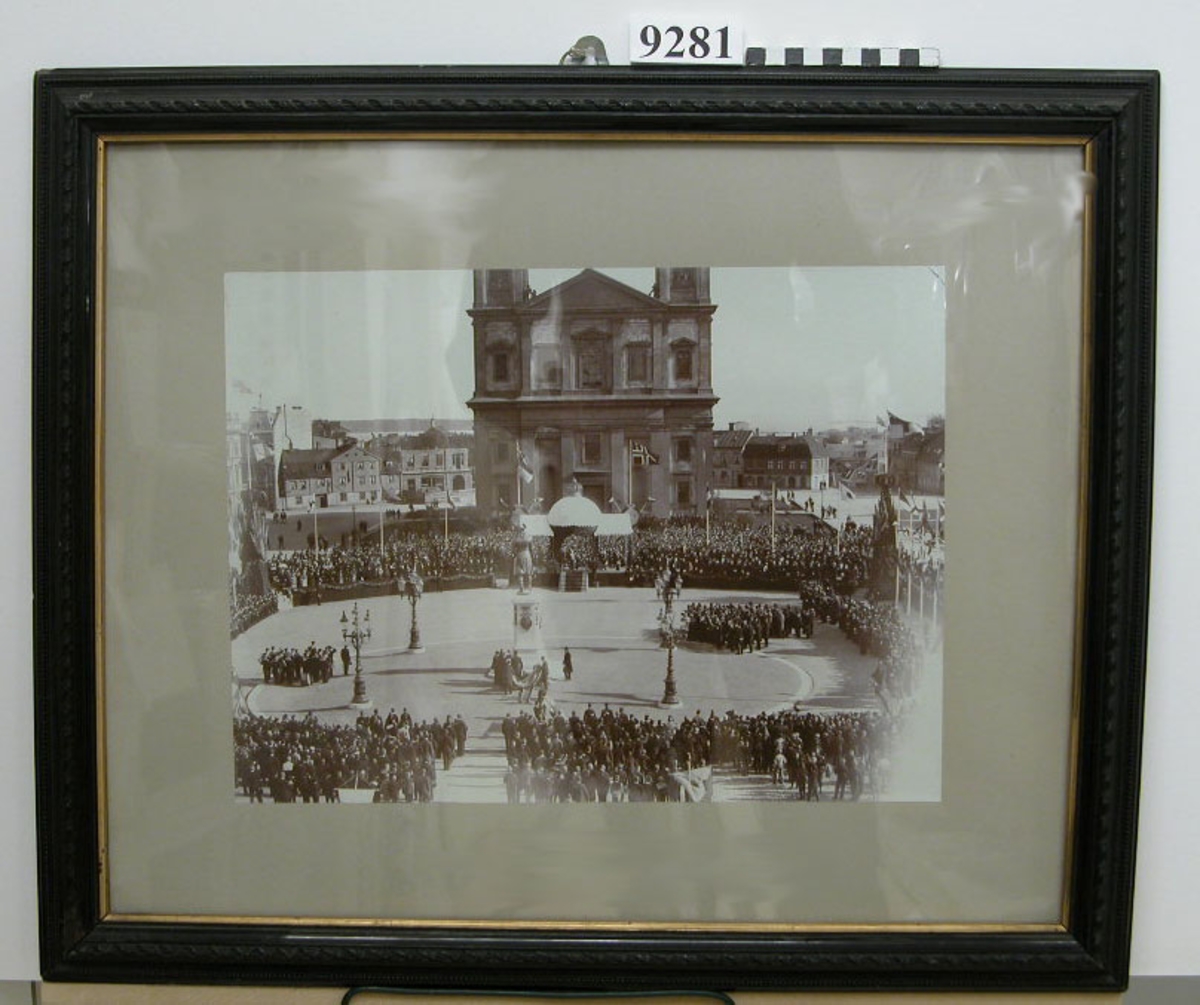 Fotografi inom glas och ram. Ramen av trä, svart med förgylld innerkant.
Föreställer avtäckningen av Carl XI:e statyn å Karlskrona stortorg år 1897.
(Dubblett finnes K 9152)