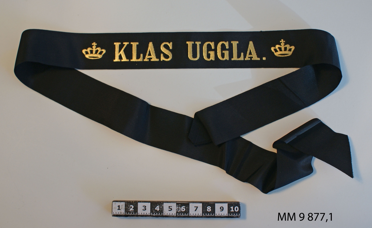 Mössband till rundmössa för manskap vid flottan.  Av svart rips med namn "Klas Uggla" i guldbokstäver, flankerat av två kungl. kronor i guld. 3 st.