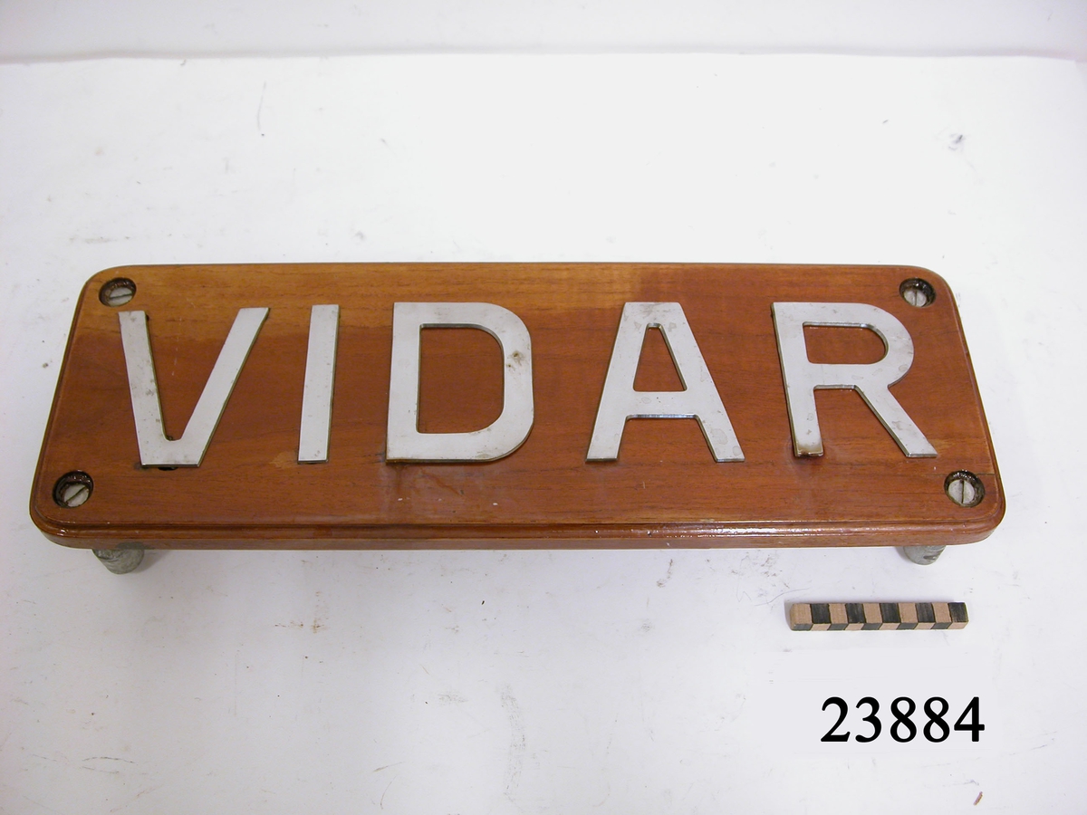 Rektangulär, lackad träplatta med rundade hörn och falsad kant. På framsidan versala bokstäver av rostfritt stål "VIDAR" som är fastskruvade från brädans baksida. Bokstävernas höjd: 100 mm. Hål i plattans vardera hörn för uppmontering med fyra genomgående skruvar och muttrar.