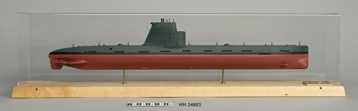 Ubåtsmodell Hajen III i monter. Modell av alträ med detaljer av mässing, målad med cellulosafärg. Rött och svart skrov. Monter av plexiglas på träplatta. Mässingsbricka i montern med uppgifter om modellen.