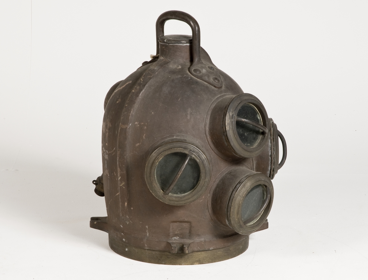 Dykarhjälm, modifierad Fahnehjelm från ca 1840-50-tal, med 4 fönster försedda med gardejärn. Upptill handtag, kort rör på baksidan för tilloppsluftslangen med backventil inuti hjälmen. Intern luftkanal från höger till vänster sida gående genom backventil med utlopp på vänstra sidan.
Från Modellkammaren.