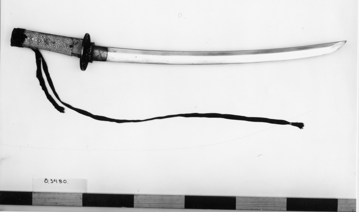 Samurajsvärd från Japan. Klinga svagt bakåtböjd, eneggad skarp, facetterad rygg. Fäste av vitt hajskinn (stingrocka?), flätad med svart bomullsgarn samt försett med två amuletter, den ena med guldinläggningar. Fästets beslag upptill och nedtill har förgyllda inläggningar. Klinga märkt: Lindman. Parerplåt rund, genombrutet bladverk med fåglar.