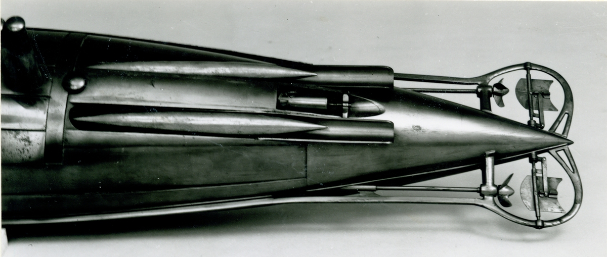 Spolformad. Stålplåt. Slingerköl, 2 propellrar av metall, 2 st höjd- och sidoroder parallellkopplade med gardjärn. 2 st höj- och sänkbara periskop av mässing. 4 st rännor för torpeder, därav 2 med luckor framtill, dels 2 för magasinering. Kort cylinder, rörlig i ränna midskepps akterut. Torpedrännorna täckas av långa stållock. Tillhörande 4 st torpeder av massiv mässing. Eneroths förslag till ubåt 1897. UndervattenstorpedFöremålets form: Spolformad.