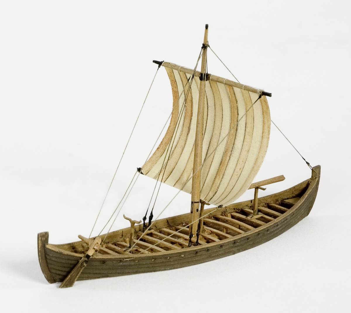 Fartygsmodell av trä. Vikingaskepp på vattenlinje. Sexton roddbänkar, styråra på styrbords sida. Riggad med en mast och ett röd-och vitrandigt segel av papper. Tältspira upplagd på bockar för om masten. Mörkbrun färg.