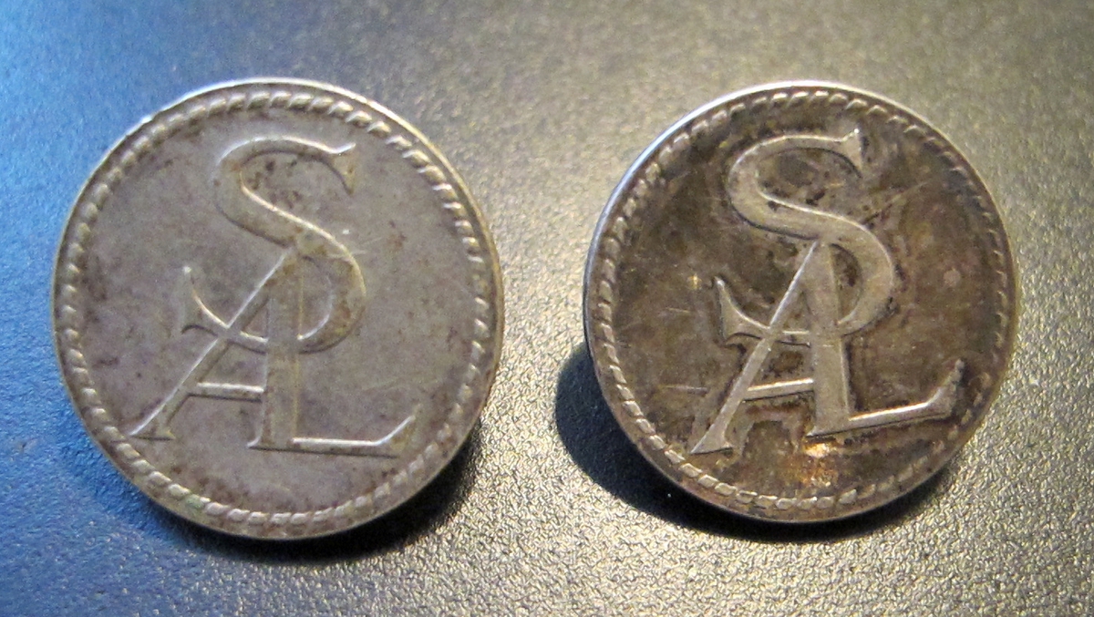 2 st cirkelrunda knappar i silver med SAL:s monogram och utmed kanten flätmönter, gjutet.

På baksidan pålödd ring för fäste på plagg, text: C.C. SPORRONG & CO STOCKHOLM
