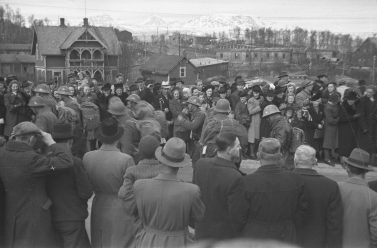 Folkemengde oppsamlet ved Narvik Jernbanestasjon. Soldater (polititropper) marsjerer fra jernbanestasjonen, 8. mai 1945. I bakgrunnen: Våningshus Rombaksveien 04. Tyske brakker