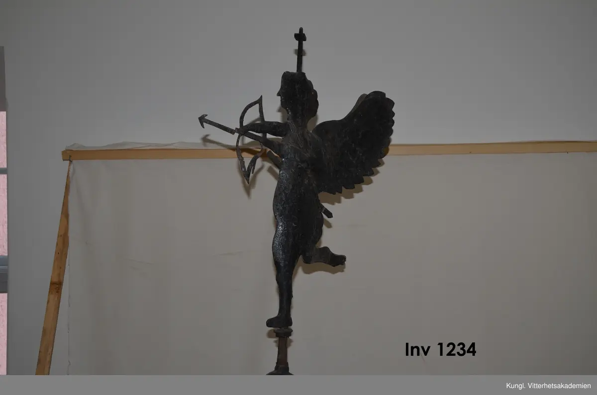Vindflöjel, i plåt, från 1700-talets slut. Påstång ovanför  det kärlliknande fundamentet  av driven plåt står en ängel eller amorin med pilbåge.  Under  vinmdflöjeln sitter smidesfundamentet som tidigare suttit på Skånelaholms slotts tak ovanför trapphust. Där rostade vindflöjeln och flyttades till ladugårdens tak. Hela ladugården brann ner 1912. Vindflöjeln räddades och finns nu kvar i rustkammare tillsammans med H Rettigs allmogesamling.