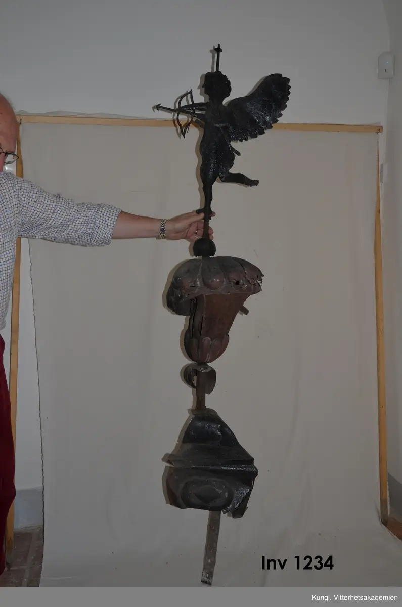 Vindflöjel, i plåt, från 1700-talets slut. Påstång ovanför  det kärlliknande fundamentet  av driven plåt står en ängel eller amorin med pilbåge.  Under  vinmdflöjeln sitter smidesfundamentet som tidigare suttit på Skånelaholms slotts tak ovanför trapphust. Där rostade vindflöjeln och flyttades till ladugårdens tak. Hela ladugården brann ner 1912. Vindflöjeln räddades och finns nu kvar i rustkammare tillsammans med H Rettigs allmogesamling.