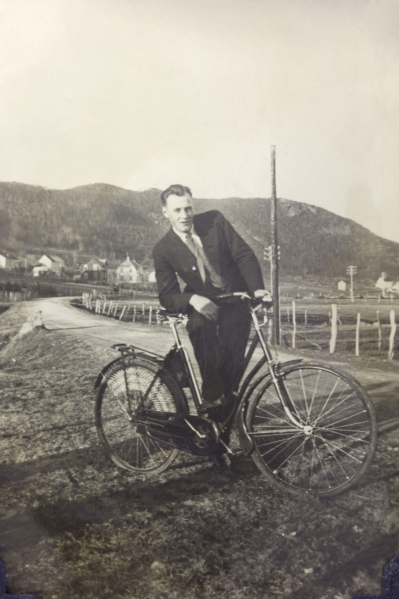 Pent kledd mann med sykkel, fotografert ved en vei. I bakgrunnen bebyggelse.